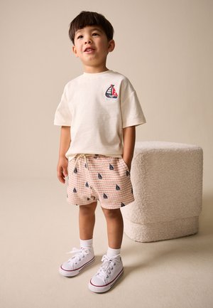 Ung dreng står iført hvid T-shirt med sejlbådslogo, mønstrede shorts med sejlbåde, hvide sokker og hvide sneakers i et neutralt rum.