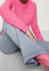 Różowy sweter z dzianiny o fakturowanym wzorze, zestawiony ze jasnoniebieskimi jeansami w dzwony. Stylizacja charakteryzuje się warstwowym wyglądem z widocznymi detalami skarpet.