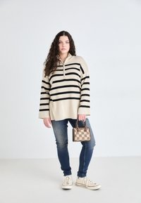 Beige striped pullover with a half-zip neck, black stripes, paired with blue jeans, cream sneakers, and a small patterned handbag.