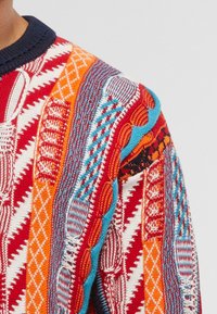Strickpullover mit einem mehrfarbigen Muster in Rot, Orange, Blau und Weiß, mit strukturierten geometrischen und wellenförmigen Designs.