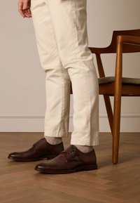 Brown leather shoes with a buckle detail, paired with light-colored pants and gray socks, set against a wooden floor and chair.