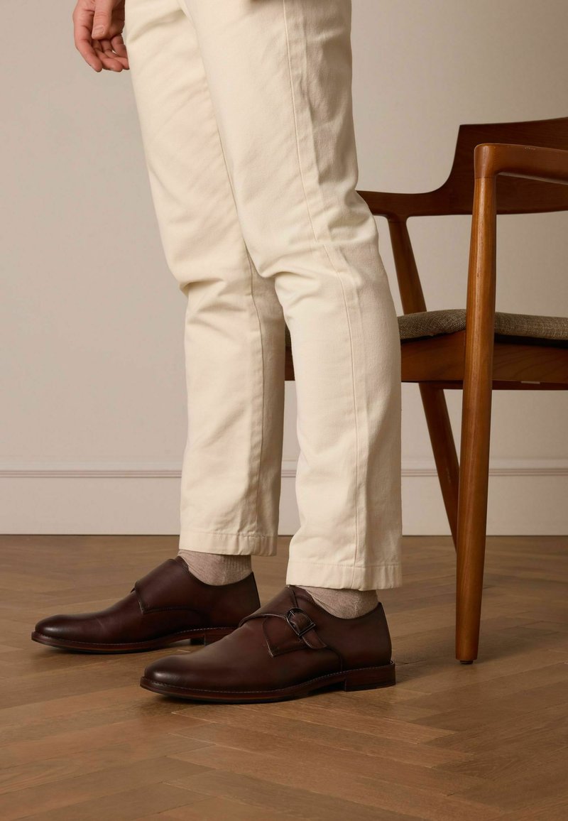 Brown leather shoes with a buckle detail, paired with light-colored pants and gray socks, set against a wooden floor and chair.