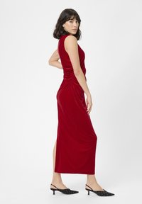 Nainen ilman hihoja punaisessa samettimekossa, jossa on sivurypytys ja halkio, sekä mustat kärjet eteenpäin suuntautuvat slingback-kengät, joissa on pieniä niittejä, poseeraa valkoista taustaa vasten.