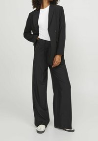 Zwarte blazer met een op maat gemaakte ontwerp, gedragen over een wit shirt. Gecombineerd met loszittende zwarte broeken en witte sneakers. Gladde textuur.