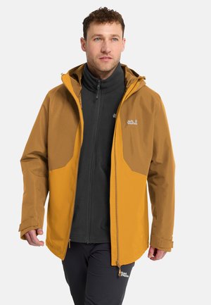 Mann trägt eine senf- und braune Outdoorjacke über einem schwarzen Fleecepullover mit Reißverschluss, mit schwarzen Hosen, steht vor einem einfarbigen hellen Hintergrund.
