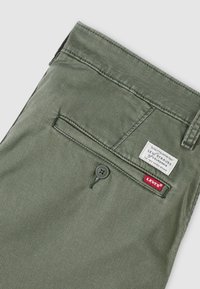 Παντελόνια Levi's σε ελιά πράσινη απόχρωση, που δείχνουν την πίσω τσέπη με κλείσιμο με κουμπί, την κόκκινη ετικέτα Levi's και την λευκή ετικέτα της μάρκας Levi Strauss.