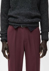 Suéter de punto acanalado gris oscuro sobre una camisa negra, combinado con pantalones burdeos de corte recto y pliegue frontal, rematados con un cinturón negro.