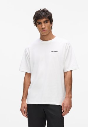 T-shirt blanc à manches courtes en coton. Il présente un col rond et un petit logo noir sur la poitrine gauche. Texture lisse, coupe décontractée.