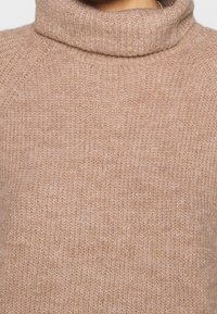 Pull en laine beige à col montant avec un tricot doux et texturé, doté d'un col et de poignets côtelés, conçu pour un ajustement près du cou.