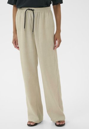 Beige wijde broek met dunne verticale strepen en een zwarte trekkoord, gedragen met een zwart topje en zwarte sandalen met open teen.