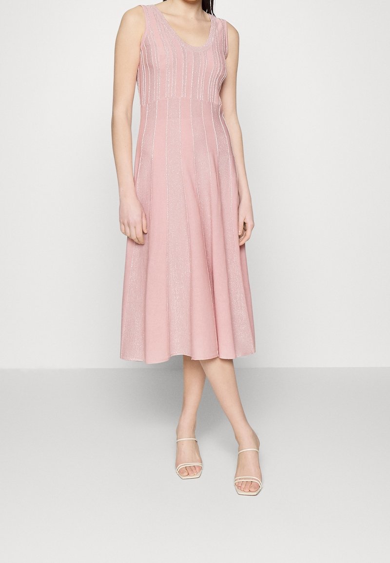 Mujer con vestido midi rosa sin mangas con rayas verticales brillantes y sandalias blancas de punta abierta, de pie frente a un fondo liso.