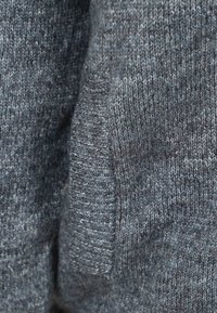 Grijze gebreide trui met een zachte textuur, subtiel patroon en geribbelde accenten aan de zoom en manchetten. Close-up van het stofdetail.