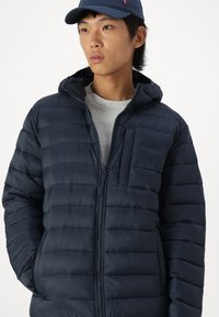 Marineblå puff jakke med horisontal søm, med glidelås foran, hette og brystlomme. Laget av lett, glatt materiale.