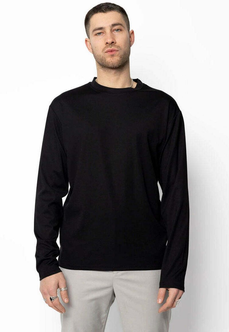 Homme portant une chemise noire unie à manches longues et un pantalon gris clair, se tenant contre un fond blanc uni.