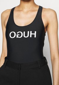 Bañador negro con tirantes anchos y escote en forma de U. Presenta un audaz logo blanco "HUGO" impreso en la parte delantera y centrado. Textura de tela suave.