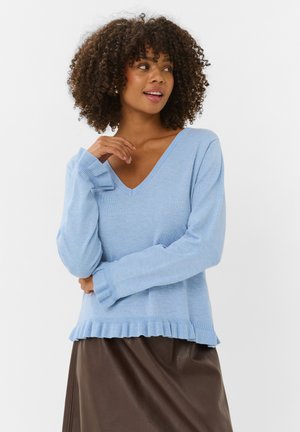 Femme aux cheveux bouclés portant un pull bleu clair à col en V et une jupe marron foncé, regardant sur le côté avec un léger sourire.