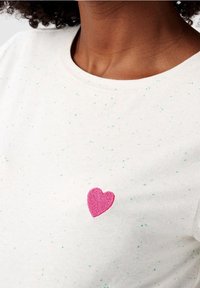 Camiseta blanca de algodón con motas verdes y azules, con un corazón rosa bordado en el área del pecho izquierdo.