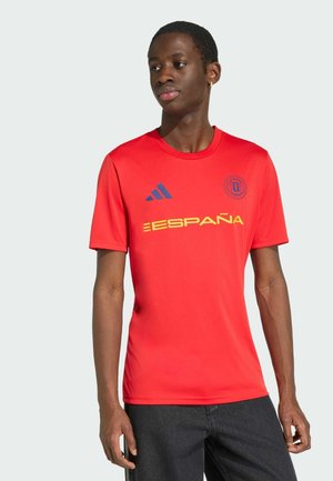 Mężczyzna w czerwonej koszulce piłkarskiej Hiszpanii z żółtym napisem „ESPAÑA”, logo Adidas i herbem drużyny, w połączeniu z ciemnymi spodniami na jednolitym tle.