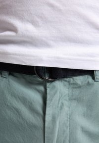 Un t-shirt en coton blanc avec un ourlet au-dessus d'une ceinture tissée noire avec un anneau en métal argenté, associé à un short texturé vert clair avec des plis à l'avant.
