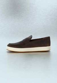 Mocassin à enfiler en cuir marron foncé avec une semelle en caoutchouc blanche, présenté sur un fond gris clair uni.