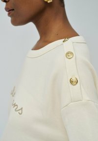 Sweatshirt crème avec un large col, accents de boutons dorés sur l'épaule et texte brodé dans une teinte marron subtile. Texture de tissu douce.