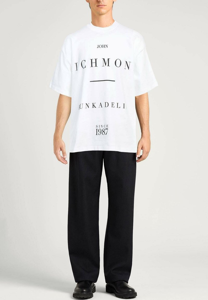 T-shirt oversized bianca con testo nero, tra cui "JOHN" e "JUNKADENI," abbinata a pantaloni larghi neri e scarpe nere.