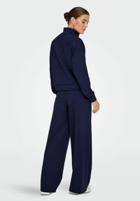 Survêtement en polaire bleu marine avec un col haut, une coupe décontractée et un pantalon large. Le matériau semble doux avec des coutures minimales et des accents épurés.