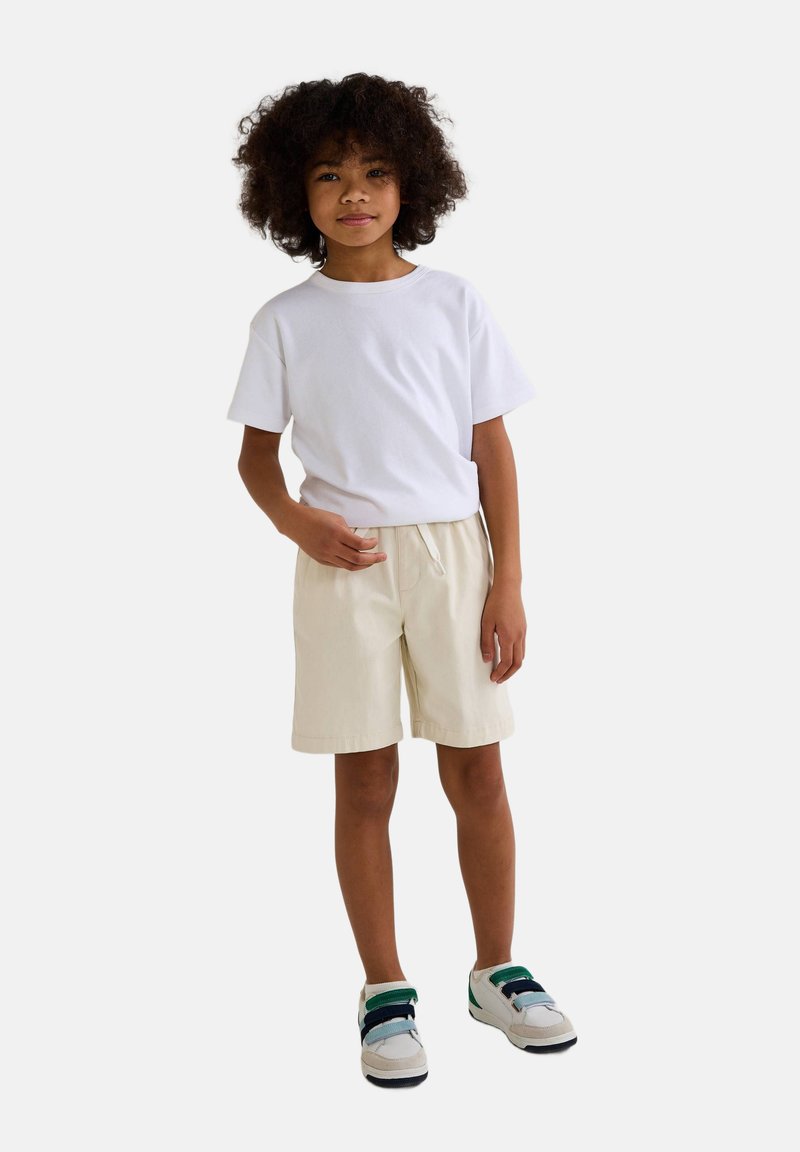 T-shirt blanc à manches courtes associé à un short beige. L'enfant porte des baskets multicolores avec des bandes Velcro. L'arrière-plan est blanc uni.