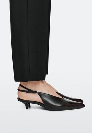 Persoon die zwarte slingback kittenhakken met puntige neus draagt, gecombineerd met zwarte broek met rechte pijpen tegen een effen achtergrond.