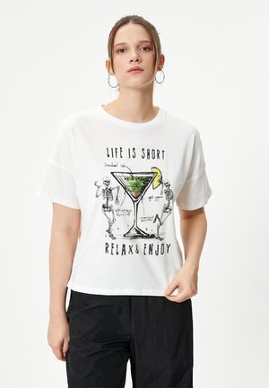 T-shirt blanc à manches courtes avec un graphique de deux squelettes à côté d'un verre à cocktail contenant un liquide vert et une tranche de citron jaune. Le texte indique "LA VIE EST COURTE, DÉTENDS-TOI ET PROFITE."