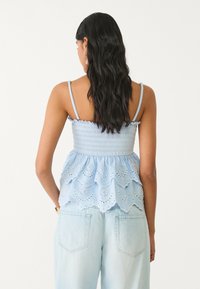 Svetlomodrý, bezrukávový peplum top s smockovaným telom a vrstveným riaseným lemom. Nosený s svetlo denimovými nohavicami, pohľad zozadu.