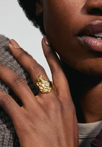 Anillo de oro con una forma orgánica, que presenta una superficie texturizada y una piedra preciosa clara, llevado en una mano descansando sobre una tela de punto gris.