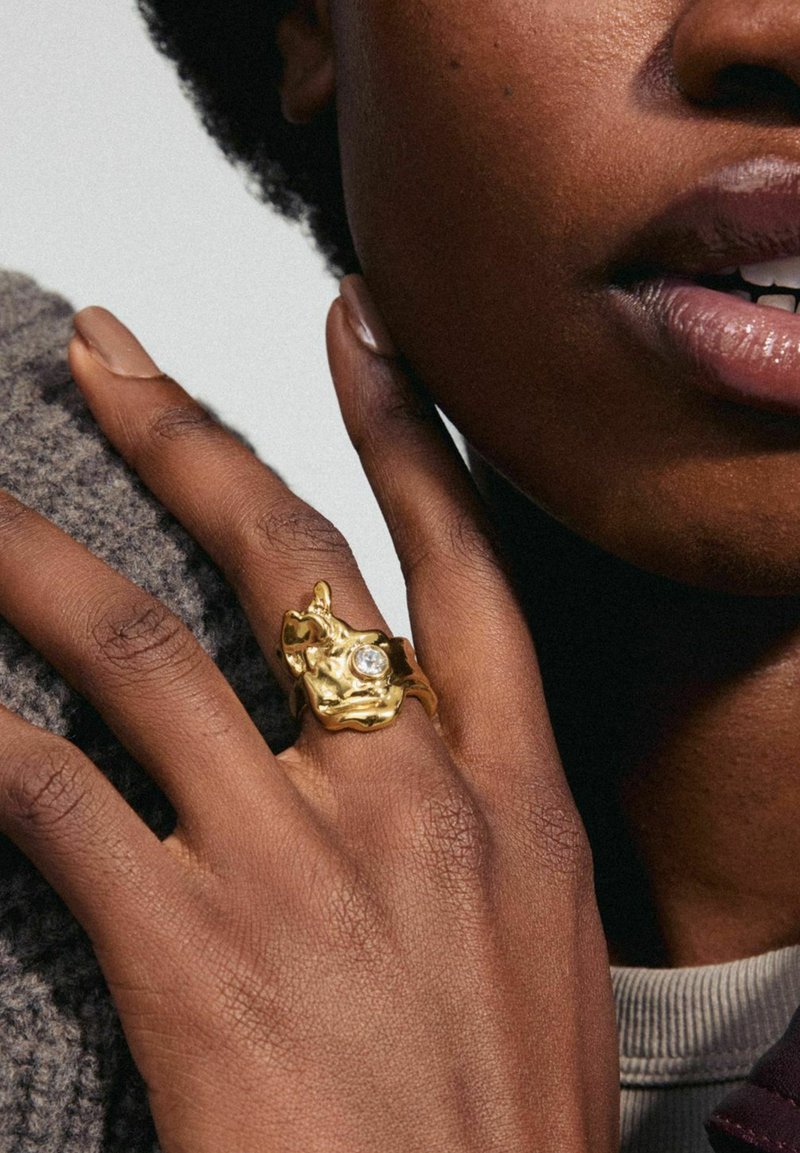 Anillo de oro con una forma orgánica, que presenta una superficie texturizada y una piedra preciosa clara, llevado en una mano descansando sobre una tela de punto gris.