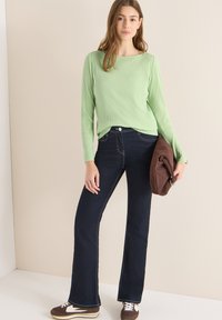 Jasnozielona bluzka z długim rękawem o prążkowanej fakturze w połączeniu z ciemnoniebieskimi, rozszerzanymi jeansami. Dodatki to brązowa torba zamszowa i brązowe trampki.