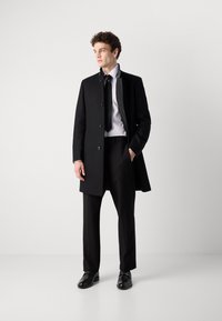 Palton negru cu două rânduri de nasturi, cu o textură netedă, asortat cu o cămașă albă și o cravată neagră. Pantaloni negri slim-fit și pantofi negri completează ținuta.