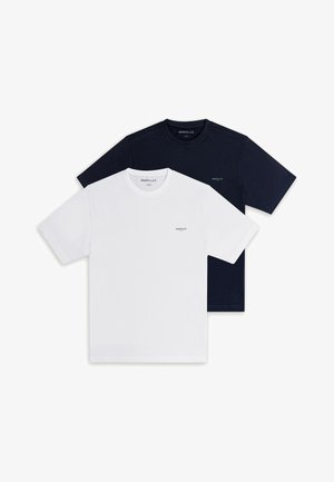 Mężczyzna w czarnych okularach przeciwsłonecznych, granatowej koszulce z krótkim rękawem, wyblakłych niebieskich dżinsach i brązowych butach, stojący z jedną ręką w kieszeni.