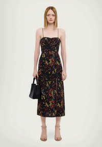 Rochie neagră midi cu bretele subțiri, cu model floral în roșu, galben și alb, având o deschidere frontală. Modelul ține o mică geantă de mână neagră.