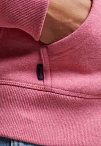 Sudadera rosa con textura acanalada, bolsillo canguro y etiqueta azul. Detalles de costura visibles en los puños y el dobladillo.