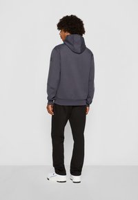Felpa con cappuccio grigio scuro dal finish testurizzato, abbinata a pantaloni neri e sneakers bianche, che mostrano un fit rilassato e uno stile casual.