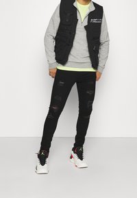 Jeans negros desgastados con rasgaduras, sudadera gris superpuesta sobre una camiseta verde neón y zapatillas negras y blancas con acentos rojos. Chaleco negro con bolsillos.