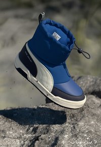 Botte imperméable bleue avec un dessus doux, un col rembourré et une semelle de couleur crème contrastante. Elle est dotée d'une fermeture à cordon et de matériaux texturés.