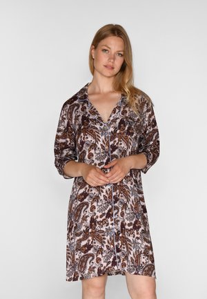 Vrouw draagt een knielange, lange mouwen, bloemenpatroon blousejurk met kraag, en staat voor een effen lichte achtergrond.