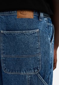 Overoles de denim azul con un gran bolsillo trasero, costuras blancas en contraste y una etiqueta de cuero marrón con un logo y texto en relieve.