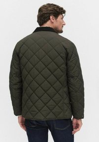 Man som står med ryggen mot kameran, iförd en mörkgrön vadderad jacka med svart krage och mörkblå jeans, mot en enfärgad bakgrund.