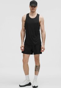 Uomo che indossa una canotta atletica nera, pantaloni corti neri, cappellino nero, calzini bianchi e sneakers bianche, in piedi contro uno sfondo neutro.