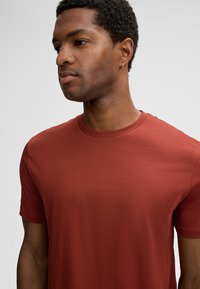 Man met kort krullend haar, gekleed in een roestkleurig crewneck t-shirt en een kleine gouden ring oorbel, kijkt naar links tegen een effen achtergrond.