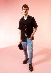 Zwarte korte mouwen shirt, lichtblauwe spijkerbroek en zwarte platform schoenen. Model houdt een zwarte pet vast. De achtergrond is een zachte roze gradient.