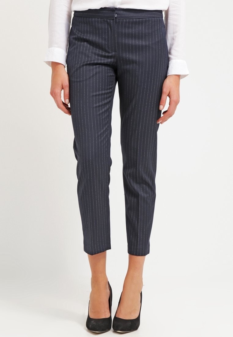 Femme portant un pantalon slim bleu marine à fines rayures aux chevilles, associé à des talons noirs et une chemise blanche manches longues.