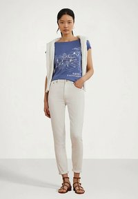 Nő kék grafikai pólóban, fehér nadrágban, bézs kötött mellénnyel a vállán, és barna pántos szandálban, fehér háttér előtt.