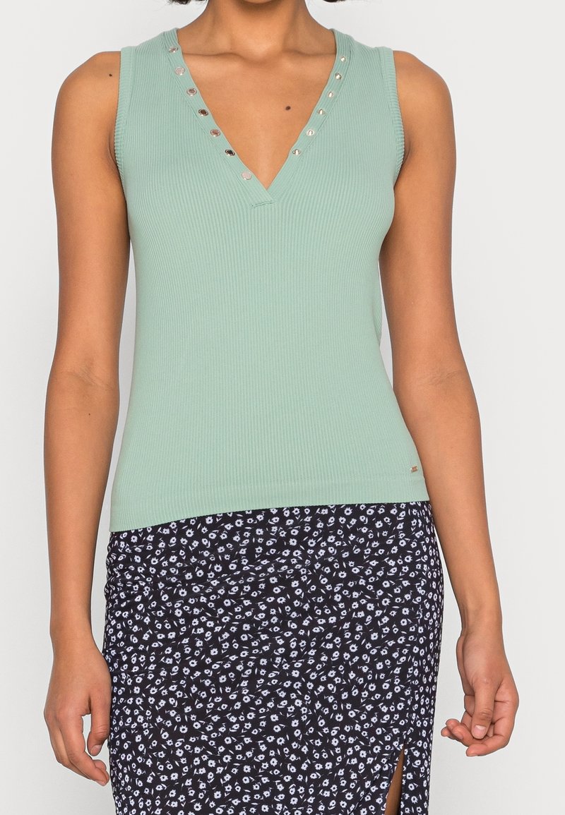 Haut sans manches côtelé vert clair avec un décolleté en V profond, ornée de boutons en argent décoratifs le long de l'encolure. Porté avec une jupe noire à motifs.